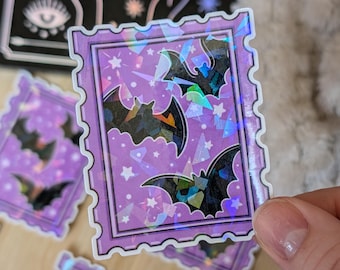 Könnte beinhalten: Ein rechteckiger Aufkleber in Form einer Briefmarke mit violettem Hintergrund, weißen Sternen und drei schwarzen Fledermaussilhouetten. Der Aufkleber hat eine holografische Oberfläche, die das Licht in verschiedenen Farben reflektiert.