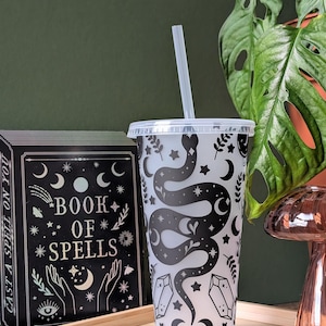 K&ouml;nnte beinhalten: Ein durchsichtiger Kunststoffbecher mit einem schwarzen Schlangen- und Himmelsdesign, einschlie&szlig;lich Monden, Sternen und Kristallen. Der Becher hat einen durchsichtigen Deckel und Strohhalm. Ein schwarzes Buch mit den Worten "Book of Spells" ist sichtbar.
