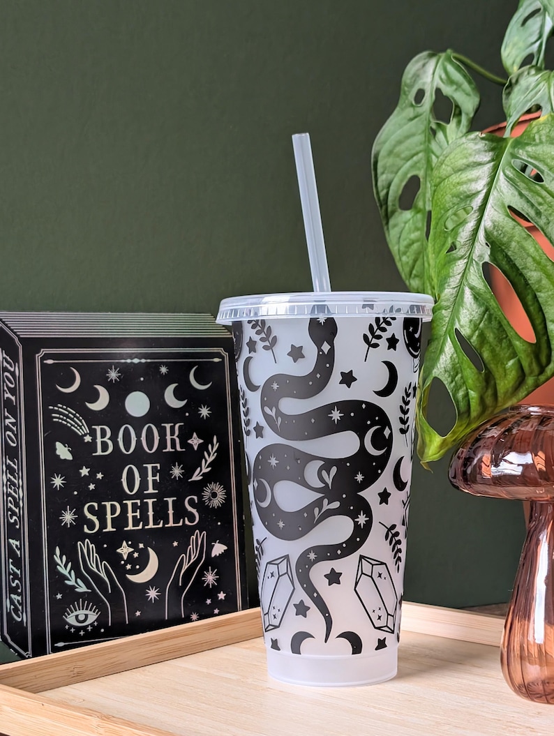 K&ouml;nnte beinhalten: Ein durchsichtiger Kunststoffbecher mit einem schwarzen Schlangen- und Himmelsdesign, einschlie&szlig;lich Monden, Sternen und Kristallen. Der Becher hat einen durchsichtigen Deckel und Strohhalm. Ein schwarzes Buch mit den Worten "Book of Spells" ist sichtbar.