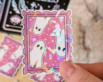 Könnte beinhalten: Ein rosa Aufkleber mit einem stempelartigen Rand, der drei weiße Geisterillustrationen mit schwarzen Augen zeigt. Der Aufkleber hat ein holografisches, irisierendes Finish und ist mit kleinen rosa Herzen verziert. Der Text "OF SPELLS" ist im Hintergrund sichtbar.