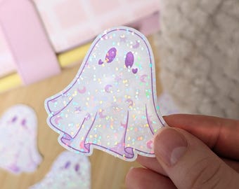 Könnte beinhalten: Ein holografischer Geisteraufkleber mit lila Akzenten, einschließlich Augen, Herzen und Monden. Der Aufkleber hat ein glitzerndes, schillerndes Finish. Zwei weitere Geisteraufkleber sind im Hintergrund zu sehen, zusammen mit einem rosa Notizbuch.
