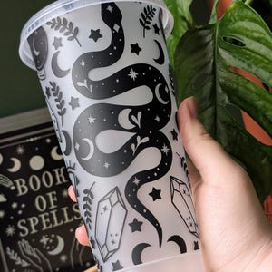 K&ouml;nnte beinhalten: Ein durchsichtiger Kunststoffbecher mit einem schwarzen Schlangen- und Himmelsdesign. Der Becher hat einen Deckel und einen Strohhalm. Das Design umfasst Sterne, Monde, Kristalle und botanische Elemente. Die Worte "BOOK OF SPELLS" sind auf einem Buch in der N&auml;he zu sehen.