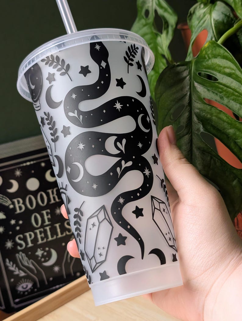 K&ouml;nnte beinhalten: Ein durchsichtiger Kunststoffbecher mit einem schwarzen Schlangen- und Himmelsdesign. Der Becher hat einen Deckel und einen Strohhalm. Das Design umfasst Sterne, Monde, Kristalle und botanische Elemente. Die Worte "BOOK OF SPELLS" sind auf einem Buch in der N&auml;he zu sehen.