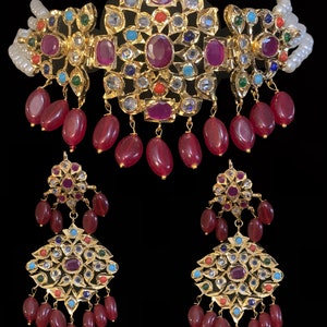 May include: A gold necklace and earrings set with intricate designs and colorful gemstones. The necklace features a large central pendant with red, blue, green, and white stones, surrounded by smaller pendants with red stones. The earrings are similar in design to the necklace, with a smaller central pendant and red stones.