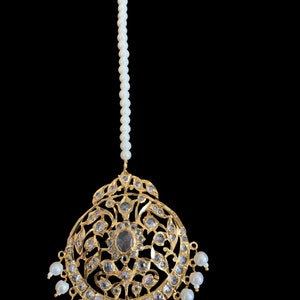 May include: A gold-toned headpiece with a central oval-shaped stone and surrounding clear stones. The ornate design features floral and leaf patterns. Small white pearls dangle from the bottom, and a strand of pearls extends upward.