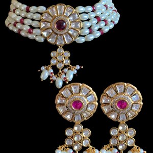 Pode incluir: Um colar choker dourado com um grande pingente floral e brincos combinando. O colar é feito de pérolas brancas e contas vermelhas. O pingente e os brincos são adornados com pedras preciosas transparentes e rosadas.