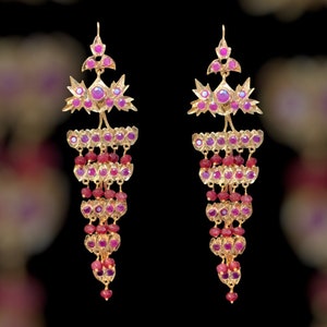 May include: A pair of gold-coloured chandelier earrings adorned with ruby-coloured gemstones. The earrings have a layered design with multiple tiers of ruby beads and larger ruby accents, creating a cascading effect. They have a hook closure.