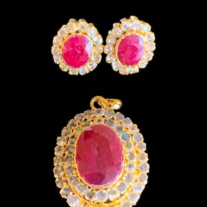 May include: A gold pendant and earrings set with a large oval ruby surrounded by smaller clear stones. The ruby is a deep red color.