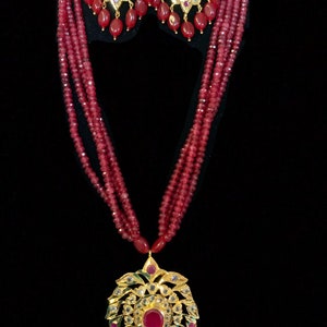 May include: A gold-toned necklace and earring set with ruby-red beads and accents. The necklace features a large pendant with a central ruby, surrounded by smaller gems and dangling ruby beads. The earrings mirror the pendant design.