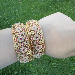 May include: Two gold-colored bangles with intricate floral patterns. The bangles are adorned with red and white gemstones, creating a striking contrast against the gold. The design is detailed and ornate, suggesting a focus on jewelry and accessories.