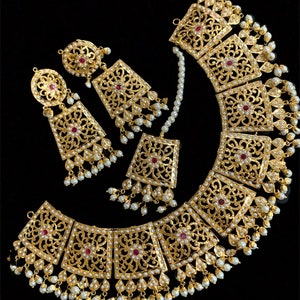 Peut inclure: Un ensemble de collier et boucles d'oreilles en or avec des motifs filigranés complexes et des accents de perles. Le collier présente une série de pendentifs de forme carrée avec des pierres précieuses rouges au centre. Les boucles d'oreilles sont d'un design assorti avec un seul pendentif carré.