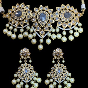 Puede incluir: Un collar de oro con tres colgantes grandes y ornamentados con piedras grises y cuentas blancas. Pendientes a juego con diseños similares y cuentas blancas.