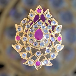 May include: A gold-colored pendant with a floral design. The pendant features a large, teardrop-shaped, magenta-colored stone in the center, surrounded by smaller, round, light blue and magenta stones. The pendant is approximately 2 inches in length.