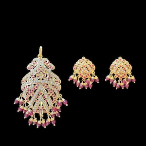 May include: A gold-toned jewelry set featuring a pendant and matching earrings. The pendant and earrings are adorned with clear and ruby-colored gemstones. The earrings and pendant have a dangling ruby-colored bead design.