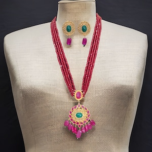 Pode incluir: Um colar de ouro com um grande pingente que apresenta uma pedra verde cercada por pedras rosa menores. O colar é feito de múltiplos fios de contas vermelhas. Brincos combinando com pedras verdes também estão incluídos.