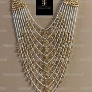 Peut inclure: Un collier multi-rangs avec des perles blanches et des accents dorés. Le collier présente un pendentif triangulaire avec des motifs floraux complexes et des pierres scintillantes.
