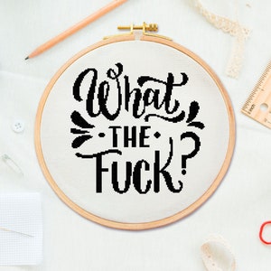 May include: A white embroidery hoop with a black cursive font saying "What the Fuck?" The hoop is on a white surface with a pencil, a ruler, a pin, and a pair of scissors.