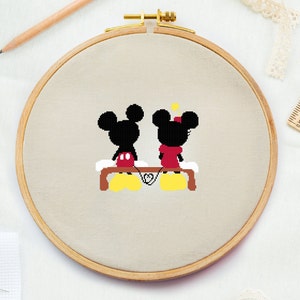 Op de afbeelding: Een borduurring met een kruissteek-borduurwerk van Mickey Mouse en Minnie Mouse die hand in hand zitten op een bankje. De achtergrond is beige.