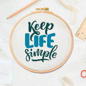May include: A white embroidery hoop with the phrase "Keep Life Simple" in black and blue lettering. The hoop is on a white surface with other craft supplies.