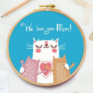 May include: Cross-stitch embroidery hoop with a blue background featuring a white cat with pink cheeks and closed eyes. The cat is surrounded by two smaller cats, one pink and one brown. The text "We love you Mom!" is written in white above the cat.