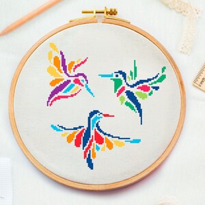 May include: A wooden embroidery hoop with three colorful hummingbird cross-stitch designs. The hummingbirds are in shades of blue, green, orange, yellow, and purple. The hoop is surrounded by craft supplies, including a pencil and ruler.