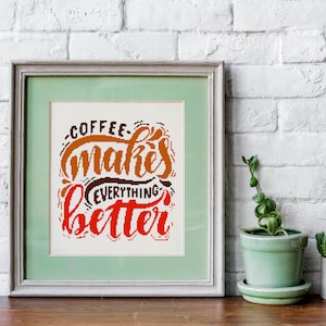May include: A framed print with a white background and the text "Coffee makes everything better" in brown, orange, and red lettering. The frame is light gray with a green mat. The print is sitting on a wooden table with two potted plants.