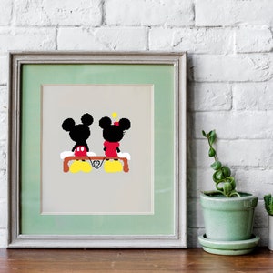 Op de afbeelding: Een ingelijst afdruk van Mickey Mouse en Minnie Mouse die op een bankje zitten en elkaars hand vasthouden. De afdruk is op een lichtbeige achtergrond met een groene passe-partout en een licht houten frame.