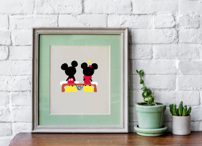 Op de afbeelding: Een ingelijst afdruk van Mickey Mouse en Minnie Mouse die op een bankje zitten en elkaars hand vasthouden. De afdruk is op een lichtbeige achtergrond met een groene passe-partout en een licht houten frame.