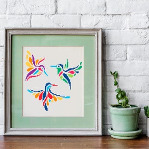 May include: Framed artwork featuring three colorful hummingbird designs in a cross-stitch style. The birds are depicted with vibrant wings in shades of red, blue, yellow, and green. The artwork is framed in a light green frame.