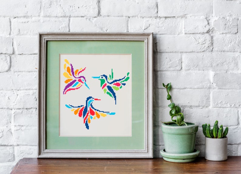 May include: Framed artwork featuring three colorful hummingbird designs in a cross-stitch style. The birds are depicted with vibrant wings in shades of red, blue, yellow, and green. The artwork is framed in a light green frame.