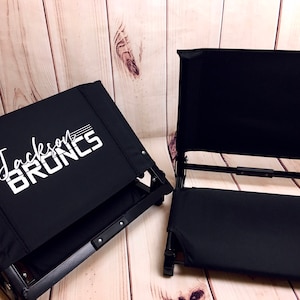Puede incluir: Dos sillas plegables negras con un dise&ntilde;o blanco. La silla de la izquierda tiene el texto "Jackson Bruins" impreso en el respaldo.