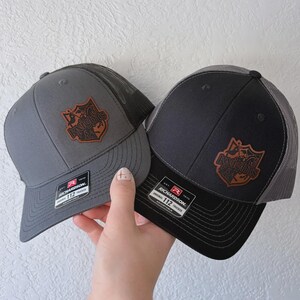 May include: Two Richardson 112 trucker hats, one gray and one black, are displayed. Each hat features a brown leather patch with the text "Knights" and a shield design. The hats have a mesh back and adjustable straps.