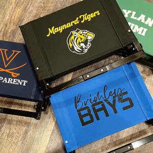 Puede incluir: Tres sillas plegables negras con diferentes logotipos de equipo. La primera silla tiene un logotipo de tigre negro y amarillo con el texto "Maynard Tigers". La segunda silla tiene el texto "Brihley Bays" en negro. La tercera silla tiene el texto "YLAN MI" en blanco.