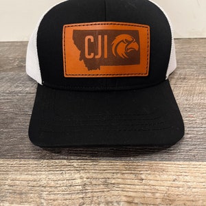 Könnte beinhalten: Eine schwarze und weiße Trucker-Kappe mit einem braunen Leder-Patch, der den Bundesstaat Montana, die Buchstaben "CJI" und einen Adler zeigt.