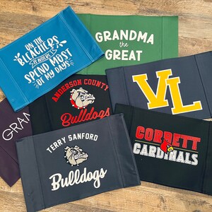 Puede incluir: Un conjunto de nueve pancartas de tela con varios dise&ntilde;os. Las pancartas presentan texto como "On the Bleachers is Where I Spend Most of My Days", "Grandma the Great", "VL", "Corbett Cardinals", "Terry Sanford Bulldogs" y "Anderson County Bulldogs". Las pancartas son de varios colores, incluyendo azul, verde, negro y gris.