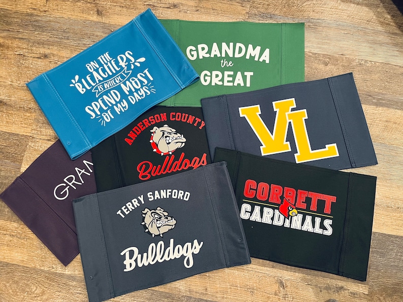 Puede incluir: Un conjunto de nueve pancartas de tela con varios dise&ntilde;os. Las pancartas presentan texto como "On the Bleachers is Where I Spend Most of My Days", "Grandma the Great", "VL", "Corbett Cardinals", "Terry Sanford Bulldogs" y "Anderson County Bulldogs". Las pancartas son de varios colores, incluyendo azul, verde, negro y gris.