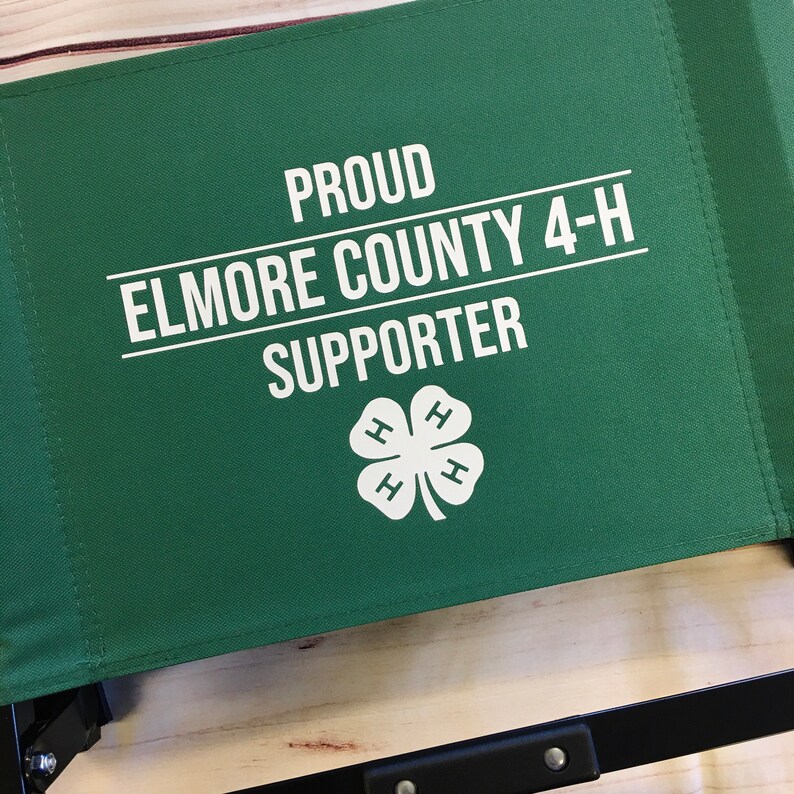 Puede incluir: Respaldo de silla de tela verde con texto blanco que dice "Proud Elmore County 4-H Supporter" y un logotipo blanco de tr&eacute;bol 4-H.