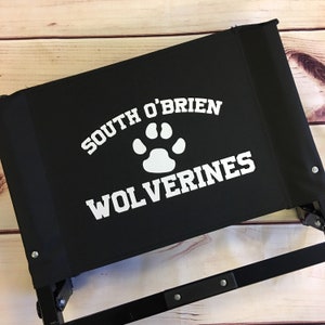 Puede incluir: Silla plegable negra con una huella de pata blanca y el texto "South O'Brien Wolverines" impreso en el asiento.