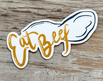 May include: A white sticker with a gold outline and the words "Eat Beef" in a stylized font. The sticker is shaped like a piece of meat.