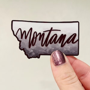 May include: A sticker featuring the outline of the state of Montana with a watercolor-style background in shades of gray. The word "Montana" is written in a stylized font in black across the state outline.