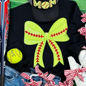 May include: Black sweatshirt with a large green and red softball bow on the front. The bow has red stitching and a white baseball design. The sweatshirt is worn over a pair of blue jeans.