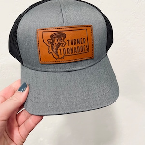 Könnte beinhalten: Eine graue und schwarze Trucker-Kappe mit einem braunen Leder-Patch, auf dem ein Tornado-Motiv und der Text "TURNER TORNADOES" abgebildet sind.