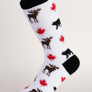 May include: White socks with a black cuff, heel, and toe. The socks feature a pattern of brown moose, black bears, and red maple leaves. The socks are displayed on a clear plastic stand.