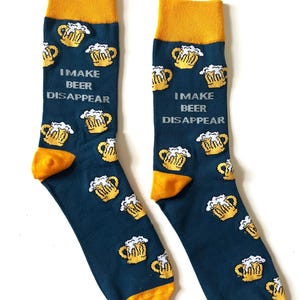 Könnte beinhalten: Ein Paar marineblaue Socken mit gelben Akzenten. Die Socken zeigen ein Muster aus gelben Bierkrügen mit weißem Schaum und dem Text "I MAKE BEER DISAPPEAR". Bündchen und Zehen sind gelb.
