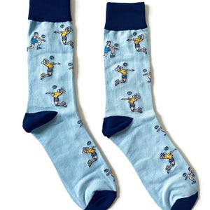 May include: A pair of light blue socks with a navy blue cuff, heel, and toe. The socks feature a repeating pattern of volleyball players in yellow and blue, mid-action, with a volleyball. These socks are a fun accessory for sports enthusiasts.