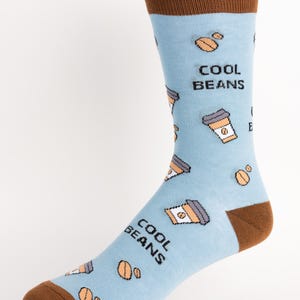 Puede incluir: Calcetines azul claro con talón, puntera y puño marrones. Los calcetines presentan un estampado repetido de tazas de café y granos de café, con las palabras "COOL BEANS" impresas en el lateral.