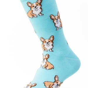 May include: A light blue sock featuring a repeating pattern of cartoon corgi dogs. The corgis are depicted in shades of brown, white, and tan, with smiling faces. The sock is made of a soft, knitted material.