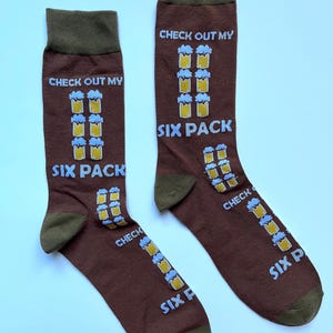 Könnte beinhalten: Ein Paar braune Socken mit grünem Bündchen. Die Socken haben einen grafischen Druck von Bierkrügen und den Text "CHECK OUT MY SIX PACK".