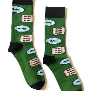 Puede incluir: Un par de calcetines verdes con talones y punteras negras. Los calcetines presentan bocadillos con la palabra "BLAH!" en blanco, rosa y azul. Un accesorio divertido y peculiar.