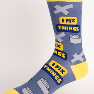 Könnte beinhalten: Eine blaue Socke mit gelben Akzenten an Zehen und Bündchen. Die Socke zeigt den Text "I FIX THINGS" in einem gelb-blauen Design, zusammen mit Bildern von Klebebandrollen und grauen Kreuzformen.